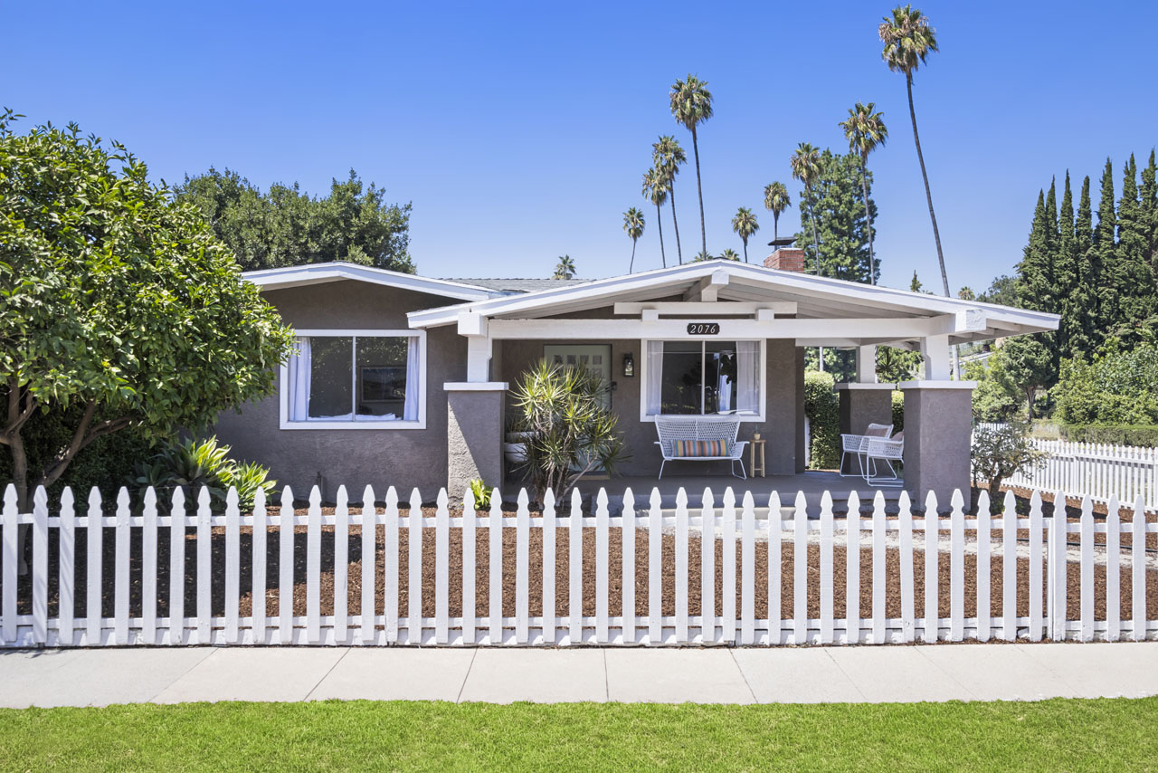 exterior of a california bungalow style home painted white and grey with a white picket fence and palm trees in the distance