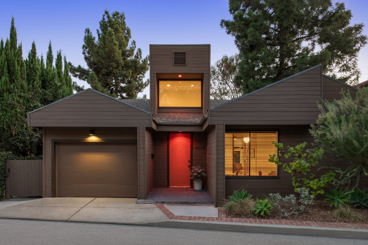 Exterior of a mid century sea ranch style home with wood siding and a red door, surrounded by greenery