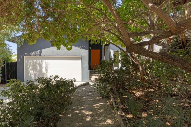exterior of a traditional home painted dark grey with a burnt orange door and front yard with a large tree and shrubs