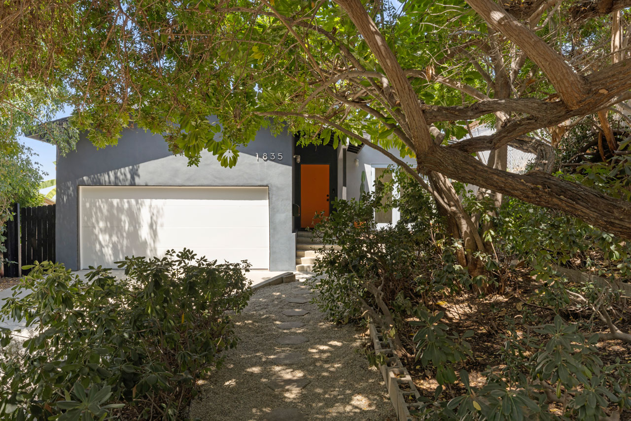 exterior of a traditional home painted dark grey with a burnt orange door and front yard with a large tree and shrubs